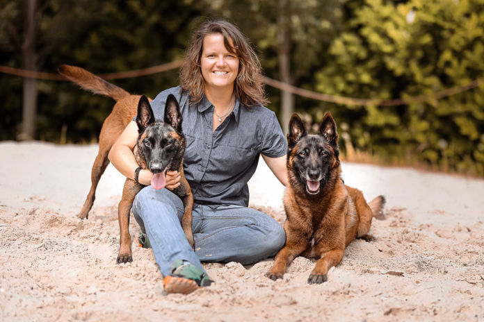 Eine Frau sitzt im hellen Sand, zu beiden Seiten ihre belgischen Schäferhunde. Einer liegt, der andere steht mit Blick zur Seite, während alle drei entspannt und freundlich in die Kamera schauen.