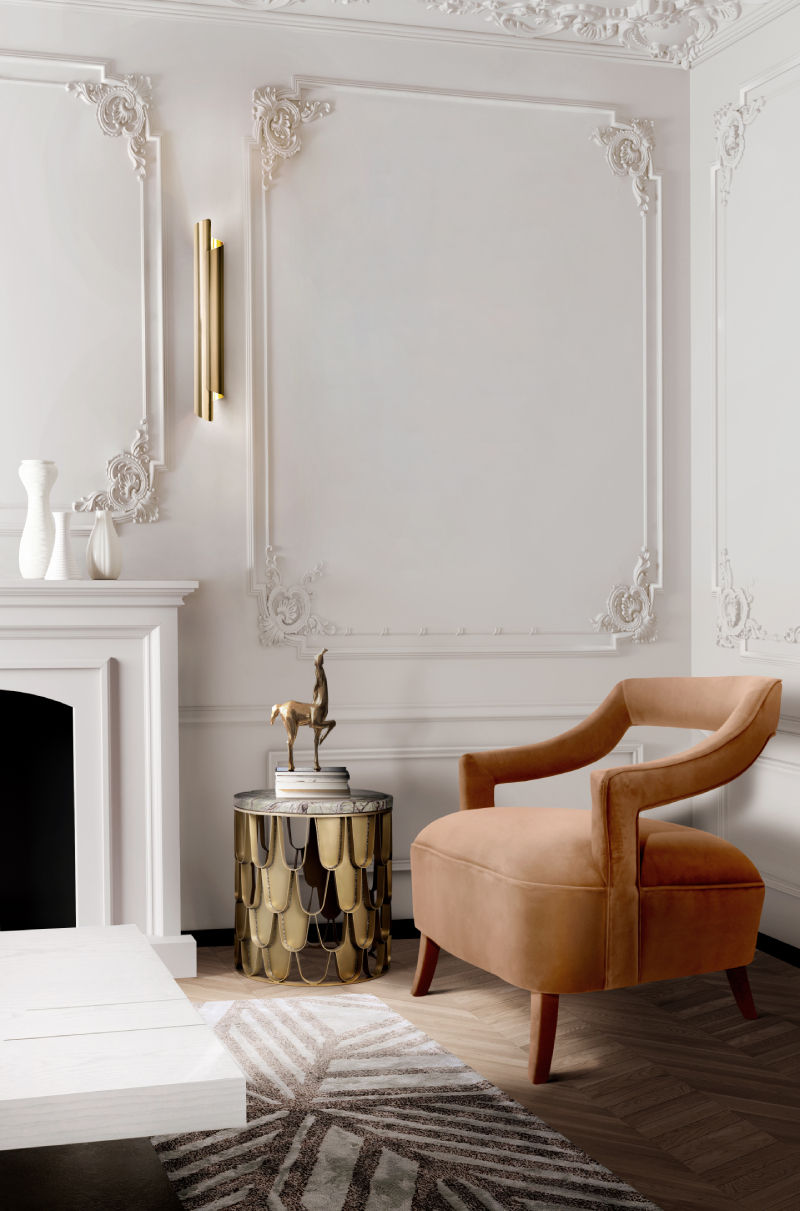 Elegant room with ornate white moldings, a tan velvet chair, a gold-accented side table, and white vases on a fireplace mantel. Cozy ambiance. The Revealry UK.