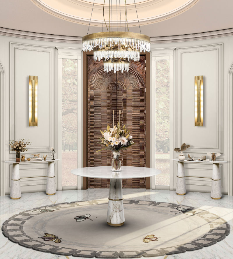 Elegant room with round white table, floral centerpiece, and crystal chandelier. Brown door, ornate rug, and gold wall sconces add luxury. The Revealry, London.