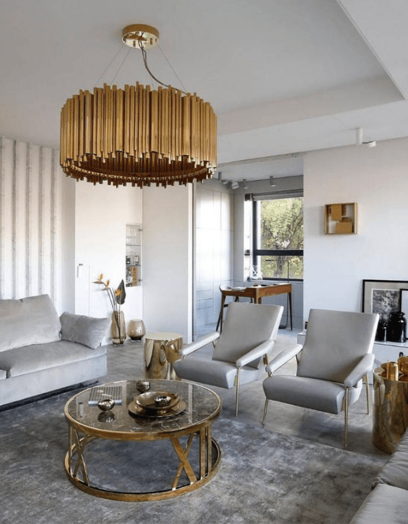 Luxurious living room with a gold chandelier, gray sofas, a round glass table, and large windows. Neutral tones create an elegant ambiance. The Revealry, London.
