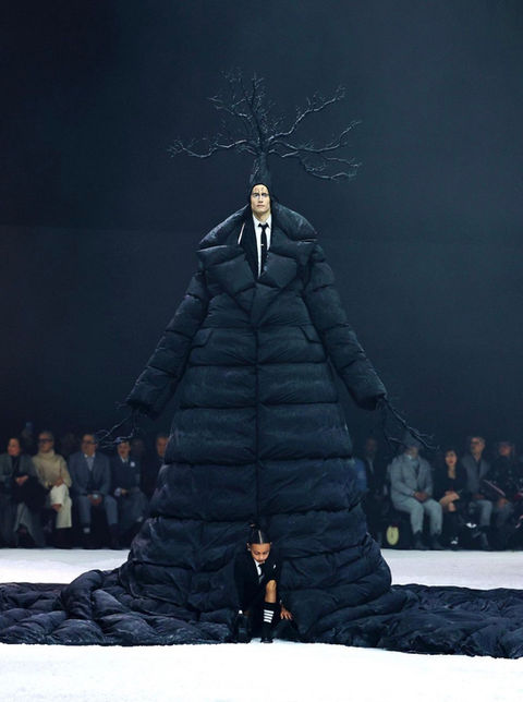 a fashion model wears an enormous puffy coat and a headpiece shaped like a tree. A child emerges from the bottom of the coat
