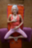 A gray-haired woman lies on her yoga mat with legs in bound angle with the soles of the feet together and the knees open wide supported by yoga bolsters. She has her right hand over her heart and her left hand on her lower abdomen. Her eyes are lightly closed and her lips slightly parted as she practices a full abdominal or diaphragmatic breath.
