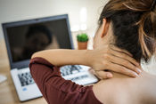 Looking over a white woman's left shoulder at an open laptop on a table in front of her. Her brunette hair is up off of her neck and she is touching her neck with her left hand as though it were stiff and painful from working on the computer.
