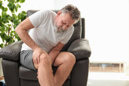 Grey haired man with full beard sits in a dark leather chair and is grasping his right leg around the knee joint with a look of discomfort on his face.