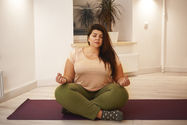 A fuller bodied woman with an olive skin tone and long brunette hair over her left shoulder sits on a yoga mat with legs crossed and arms resting on the knees holding the index finger and thumb together on each hand. Eyes are closed and there is a look of peace on her face.