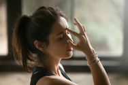 An olive complected woman with her dark brunette hair in a ponytail rests her right index finger over her third eye, her right thumb on her right nostril her right ring finger on her left nostril for directing energy and breathing in and out through alternate nostrils to balance energy in the halves of the body with alternate nostril breathing.