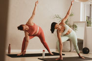 Two women practice extended side angle with their right feet pointing towards the front short edge of their mat and the right knee bent to stack vertically over the ankle bone. The left foot is about two feet behind the right with the toes pointed towards the long, left edge of their mats with the left knee straight. Their upper bodies are leaning forward towards the right knee. The right hands are touching the mat to the inside of the right foot and the left arms are extended up towards the ceiling with the palms facing the left long edge of the mat.
