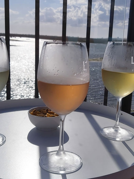 Drei Weingläser auf einem Tischchen, zwei davon mit Weißwein gefüllt, eines mit Roséwein. Im Hintergrund das Wasser einer Lagune