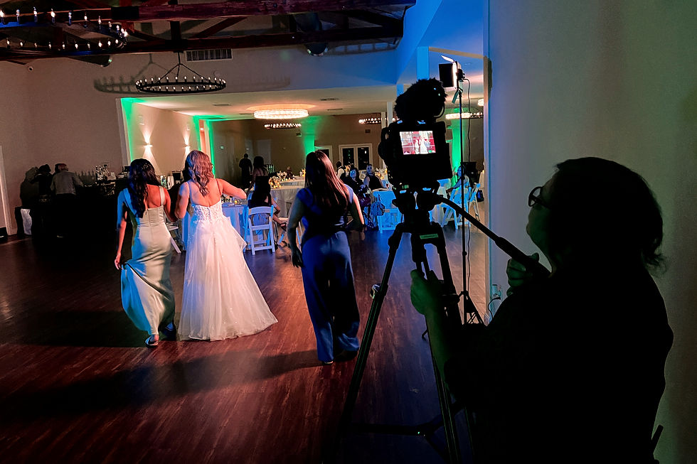 Women in formal dresses walk on a dimly lit dance floor. A person films them with a camera on a tripod. Mood is festive and lively.