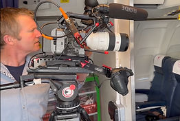 Chris, a white man with short brown hair and wearing a grey shirt, stands looking down the lens of a large video camera.