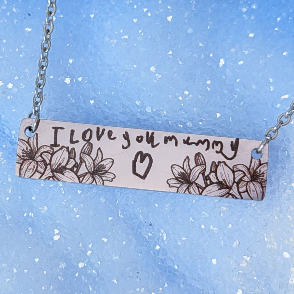 A horizontal bar necklace with an engraving of handwriting that says "I love you mummy" with a hand drawn heart and flowers.