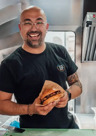 Glücklicher Mann mit Brille und Burger in der Hand, lächelnd, mit einem Tshirt wo draufsteht Foodtruck Klub