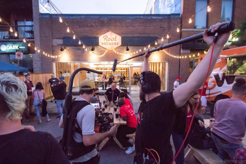Film crew recording outside "Root Brewery" at dusk. People sitting at picnic tables under string lights, vibrant, lively atmosphere.