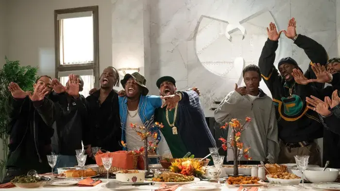 A group of people stands behind a festive table, making hand gestures and smiling. The setting features a large logo on the wall.