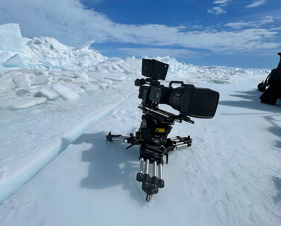 Camera on tripod on snowy terrain under a bright blue sky, surrounded by ice formations. Scene feels cold and clear.