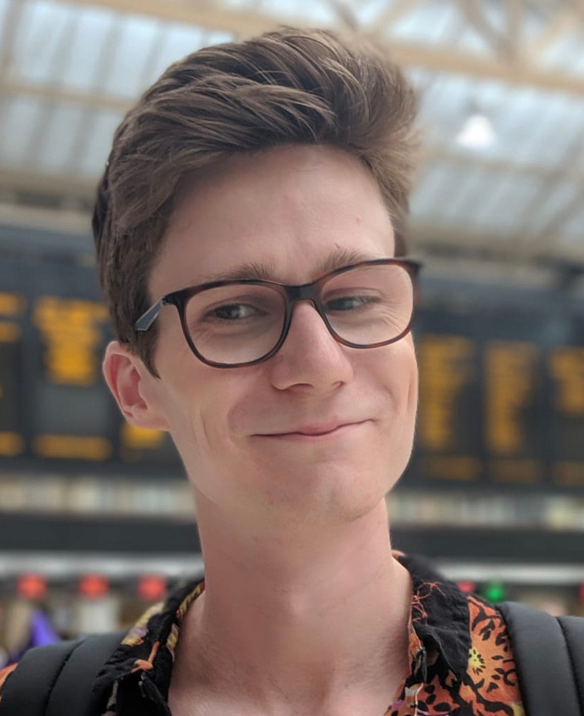A photo of Robbie, who is similar at the camera. He has light skin and short, brown hair, and is wearing glasses with black frames. The background of the image is a train station.