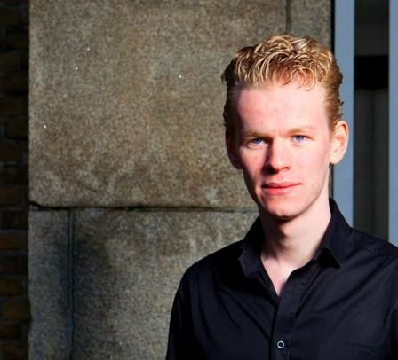 A picture of Nathan. He stands on the right side in the image, has pale skin and short blond hair. He is smiling at the camera while wearing a black shirt.