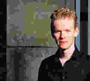 A picture of Nathan. He stands on the right side in the image, has pale skin and short blond hair. He is smiling at the camera while wearing a black shirt.