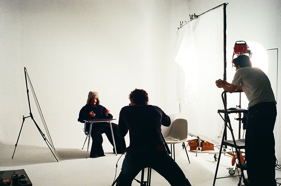 Photographer captures a person in a fur hat seated at a desk in a bright studio. Another person adjusts lighting. Minimalist setup.