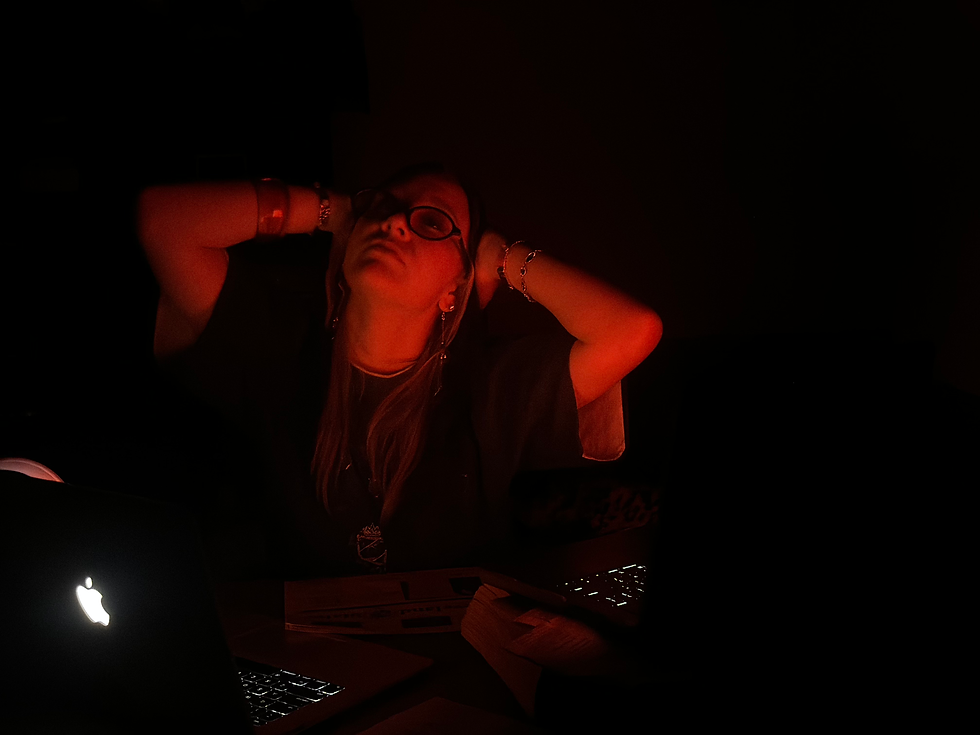 Person in a dimly lit room, leaning back with hands on head, illuminated by the glow of two open laptops. The scene is moody and introspective.