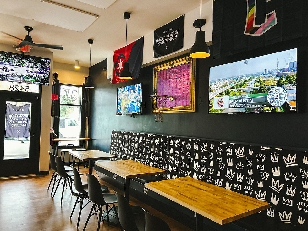 A sports bar interior with wooden tables, patterned seating, and multiple TVs showing sports. Flags and posters decorate the black walls.