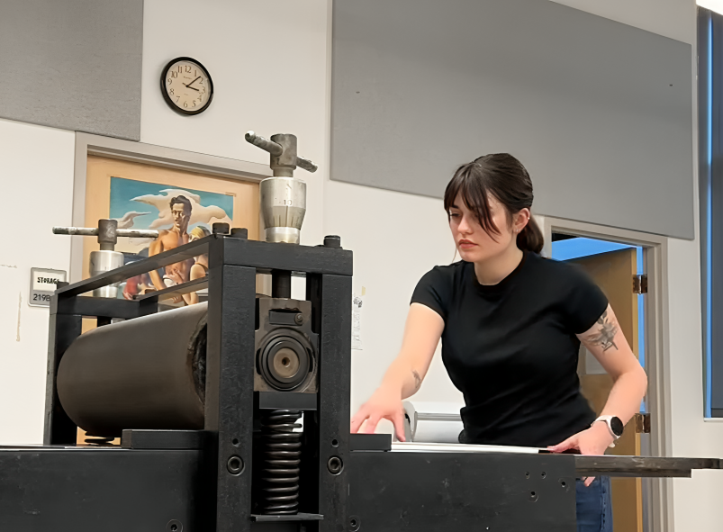 A person operates a printing press in an art studio. A clock and a painting are visible on the wall. The person appears focused.