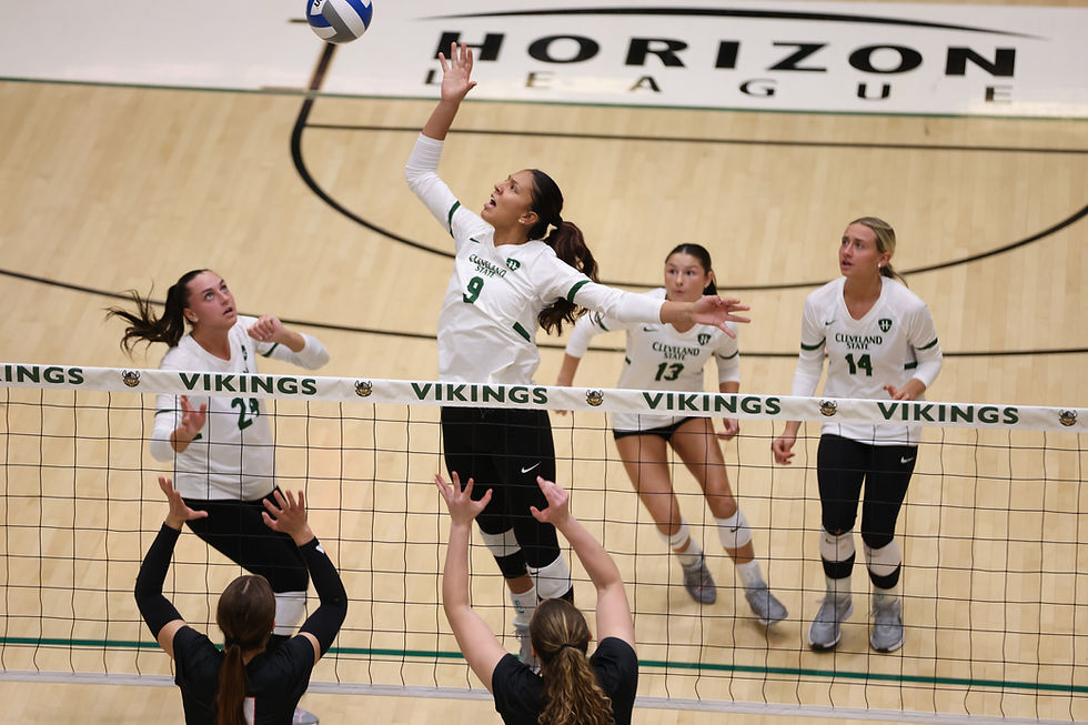 Volleyball player in white jersey jumps to spike the ball over net. Teammates and opponents watch. "Vikings" and "Horizon League" visible.