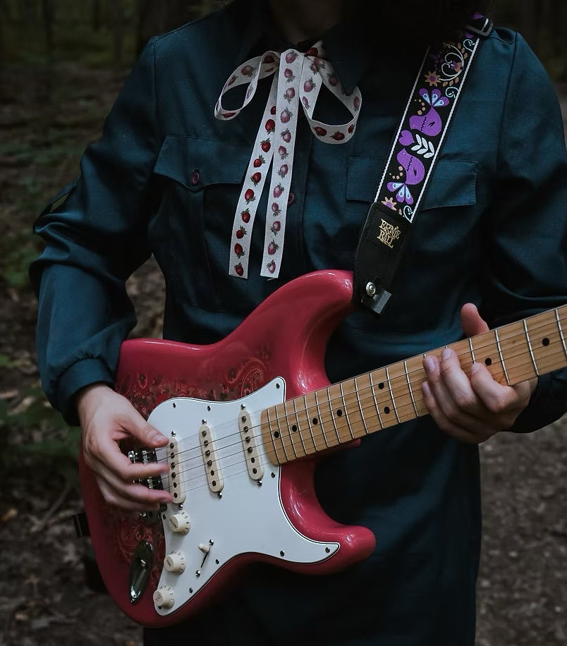 Person in dark clothing plays a pink electric guitar with a decorative strap in a wooded setting. The mood is serene and focused.