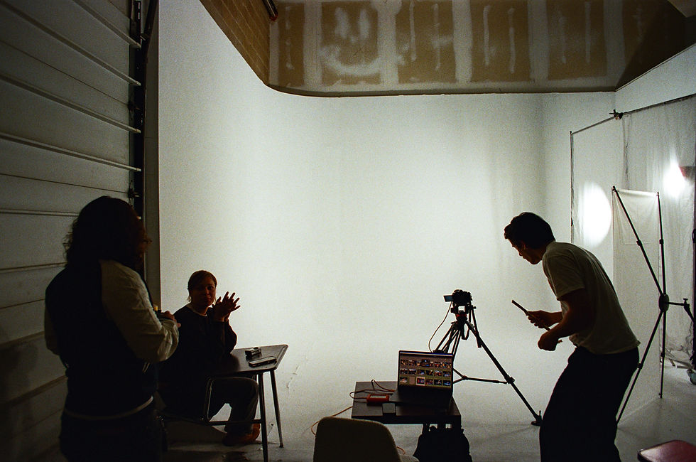 Three people in a photo studio. One adjusts a camera on a tripod, another sits at a table, and a third stands nearby. Laptop open, white backdrop.