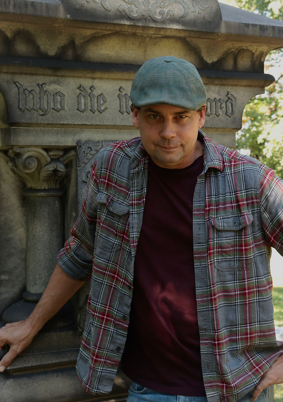 Man in a plaid shirt and cap leans on a stone monument with engraved text "who die in..." in a park. Background shows green foliage.