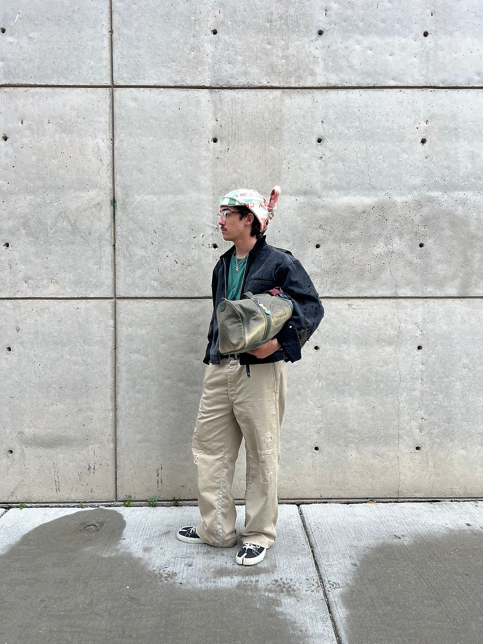 A person in a beanie and glasses stands by a concrete wall, holding a bag. They wear a dark jacket, beige pants, and casual shoes. Urban setting.
