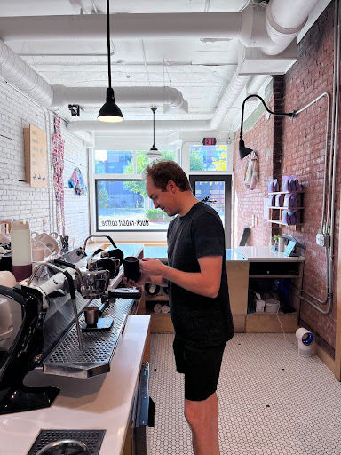 Barista in a black outfit makes coffee in a bright café with brick walls and white tiles.