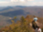 Two people on a rocky mountain peak, overlooking expansive, hazy blue hills and a partly cloudy sky. Dense green foliage surrounds them.