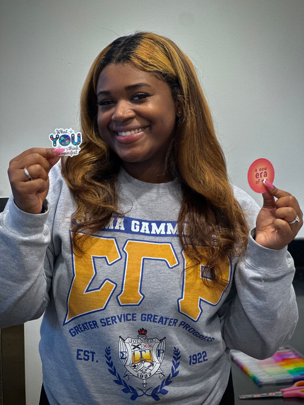 Smiling woman in a gray Sigma Gamma Rho sweatshirt holds two stickers with motivational text in a bright, indoor setting.