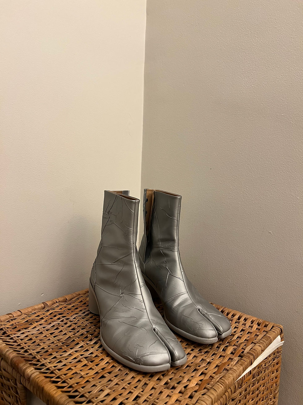 Silver, split-toe ankle boots with subtle wrinkles sit on a wicker basket against a plain, off-white wall.