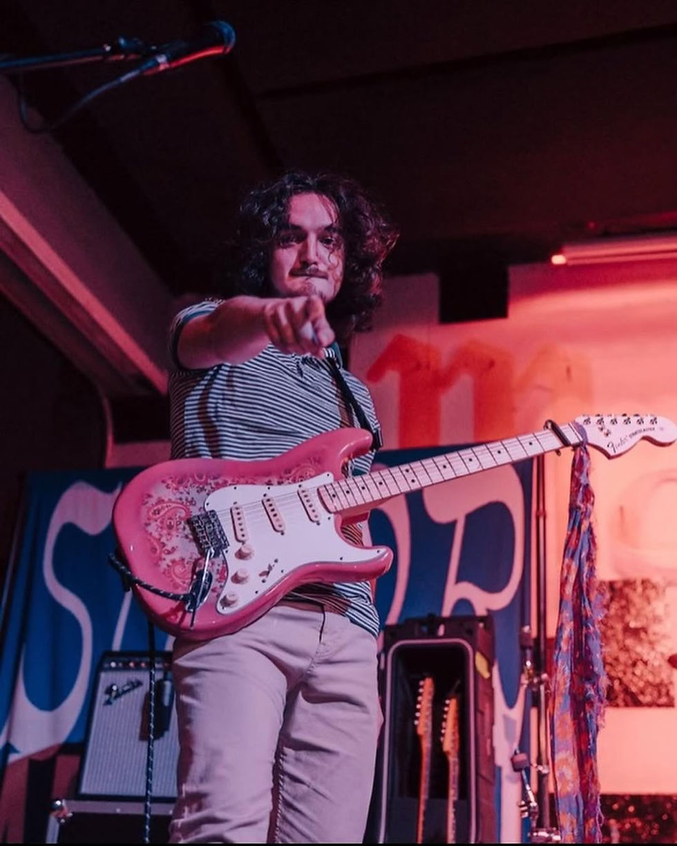 Musician with a pink guitar points at the camera on stage. He's wearing a striped shirt, with colorful lights and equipment in the background.