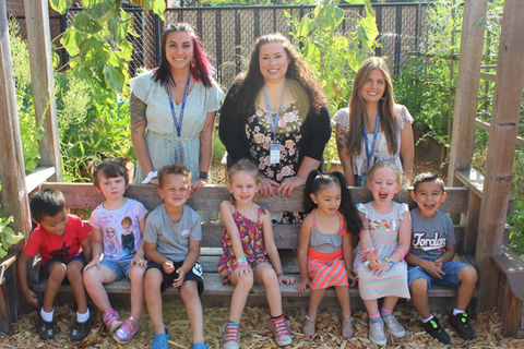 A group of seven early learners sitting on a bench outside on a sunny day with three teachers standing behind the bench above them.