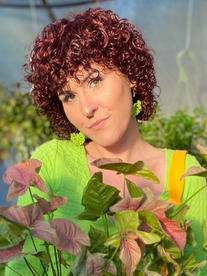 A photo of Emma Jordan, smiling with brown eyes and short red-brown curly hair. Emma has green leaf earrings, a green shirt and is holding a plant with leaves that cover her chest.
