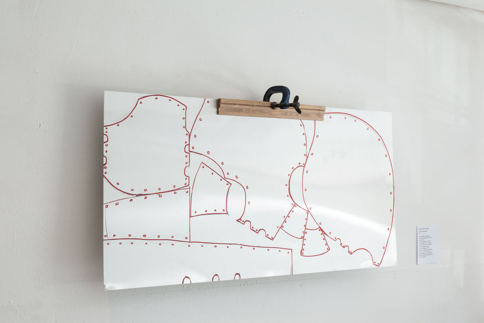A piece of white sheet metal with a dressmakers pattern drawn on in red is held to a wall in a wooden and metal clamp.