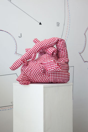 On a white plinth is a giant fabric heart, the fabric a white and red gingham pattern. In the background is a giant drawing of the fabric hearts pattern.