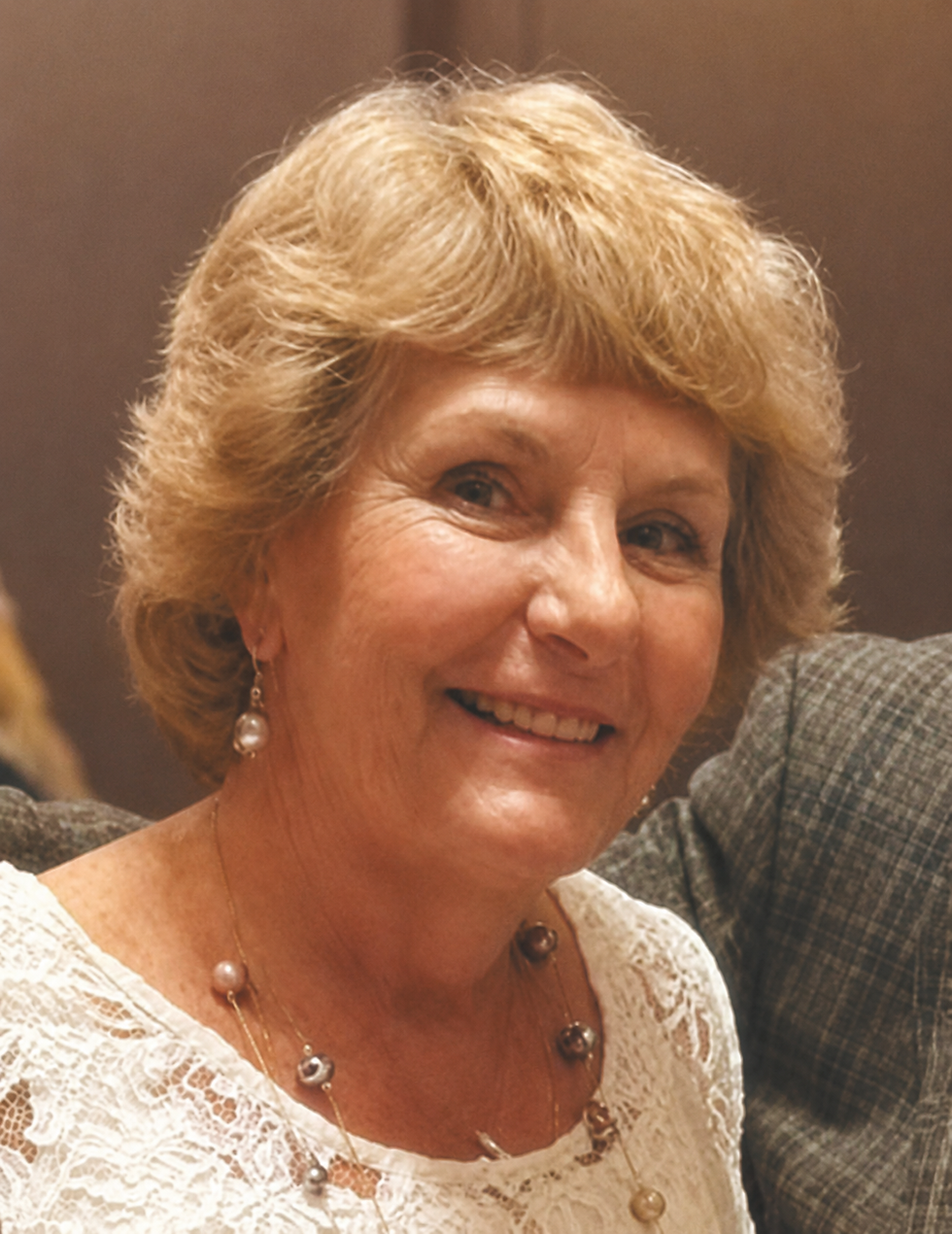 Smiling Betty Marquis with blond hair and pearl earrings in a white lace top. Warm lighting and an indistinct background.