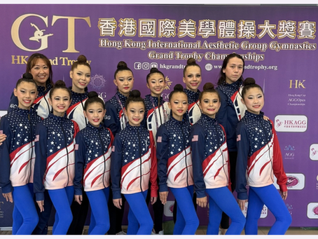 Gymnasts and accompanying adults in USA team jackets pose before a purple Hong Kong International Aesthetic Group Gymnastics competition backdrop.