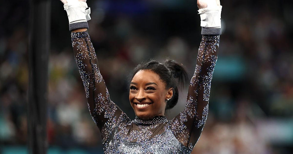 Gymnast in a sparkling leotard raises arms triumphantly, smiling in an indoor arena. Blurred audience in the background.
