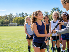 A coach welcomes a girl to field hockey practice by handing her a stick as her smiling teammates gather on a sunny field.