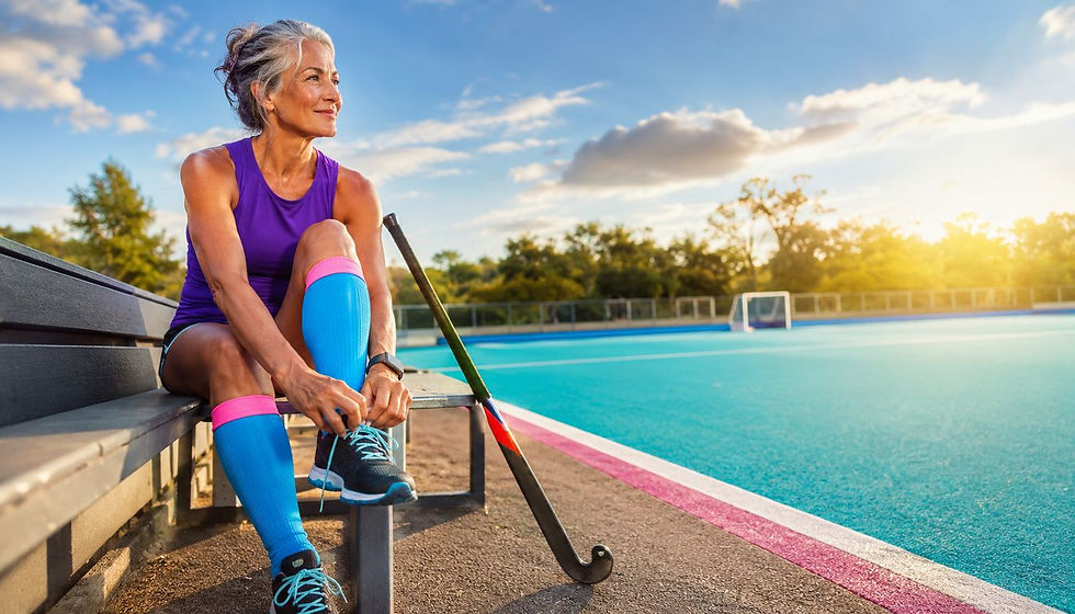 Mature female field hockey athlete resting and adjusting her shoe beside a sunlit field during recovery.