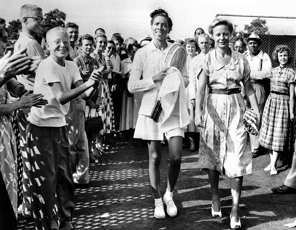 Tennis player and another person walk smiling through a cheering crowd. They're outdoors on a sunny day, holding a tennis racket and towel.