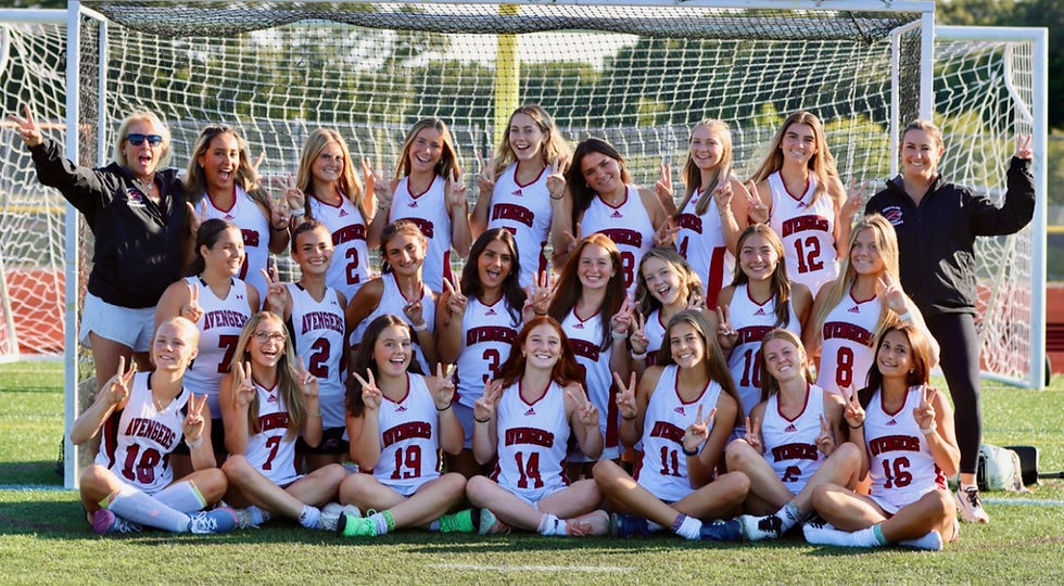 Women’s soccer team poses in front of a goal, smiling and flashing peace signs in white Avengers jerseys.