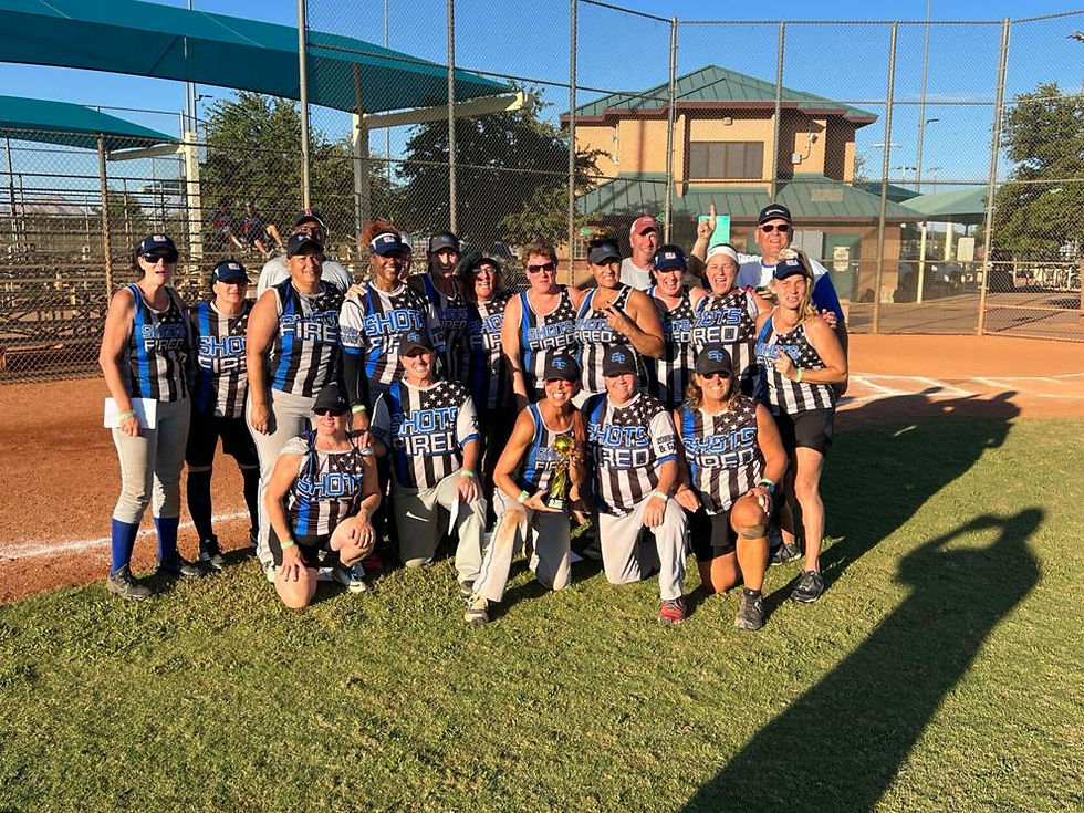 A softball team poses on a field with "SHOTS FIRED" jerseys, smiling under a sunny blue sky, near chain-link fences and a building.