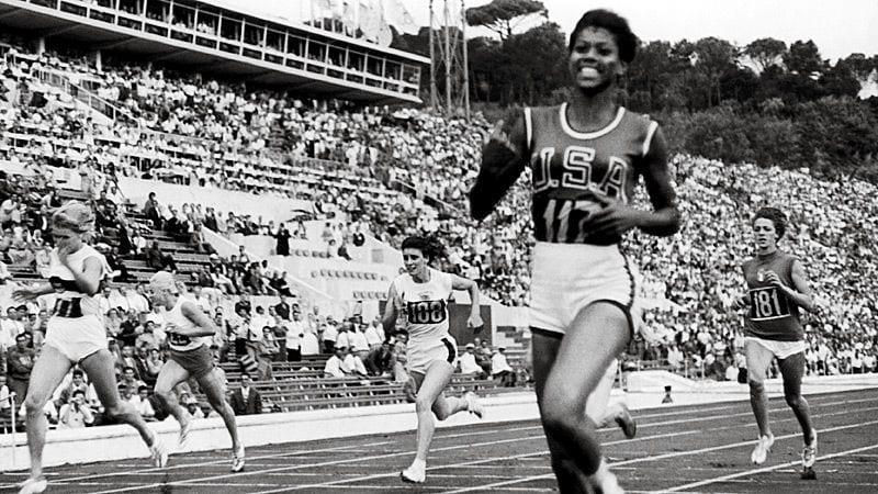 Wima Rudolph sprints on a track in a stadium, one wearing a "USA" jersey leading, smiling. Crowd in stands, trees in the background.