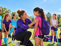A woman coach welcomes a girl to a sunny field as teammates prepare for soccer and field hockey practice.